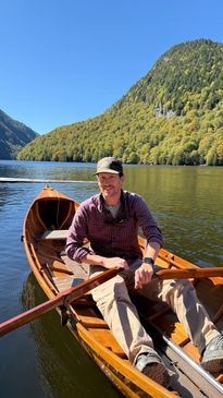 A man rows a wooden boat on a calm lake surrounded by green hills.