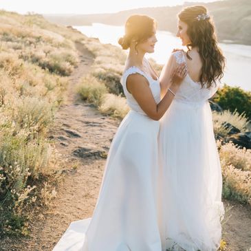 Two brides in white gowns share a moment outdoors near a river at sunset.