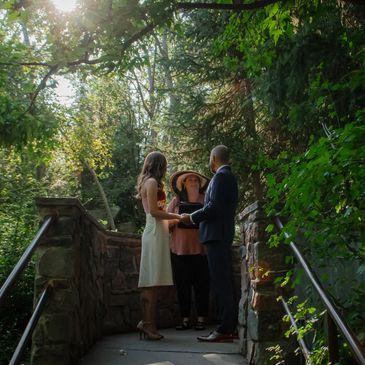 Couple exchanging vows outdoors in a sunlit, leafy setting with an officiant.