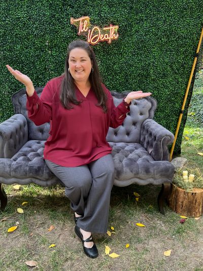 Woman in red blouse sitting on a gray tufted sofa outdoors, smiling with arms raised.