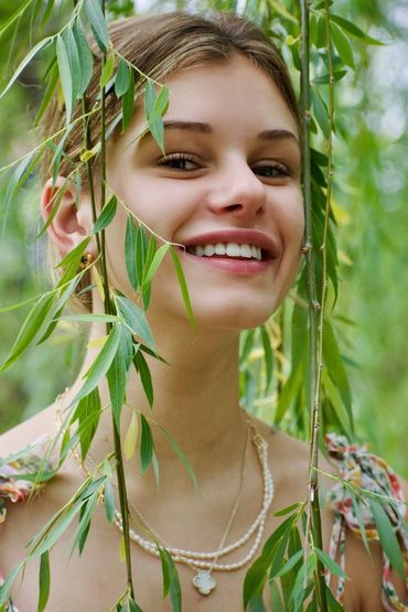 Young woman smiling behind green willow branches outdoors.