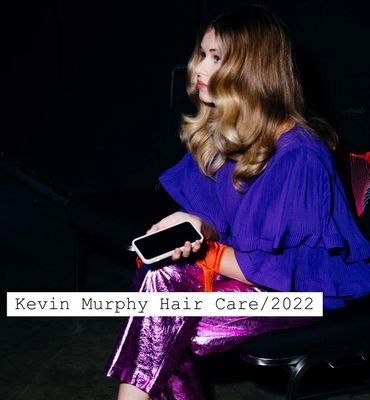 Woman with shiny styled hair wearing a purple blouse and metallic pants, holding a phone.