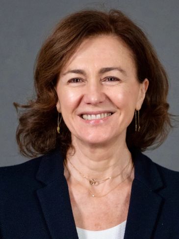 Smiling woman with brown hair wearing a navy blazer and delicate jewelry.