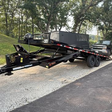 Black flatbed tilt trailer in a wooded area.