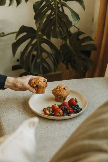 Hand holding a muffin with another muffin and fruits on a plate.