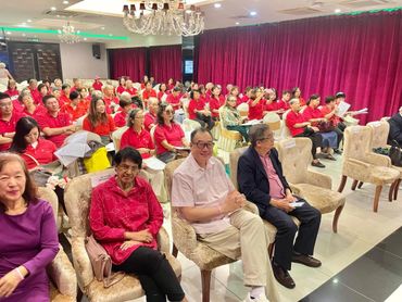 Audience in red shirts attentively seated in a conference room with chandeliers.