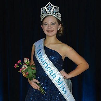 In her navy sequins gown, American Miss National Preteen smiling with her new AMP Crown and banner.