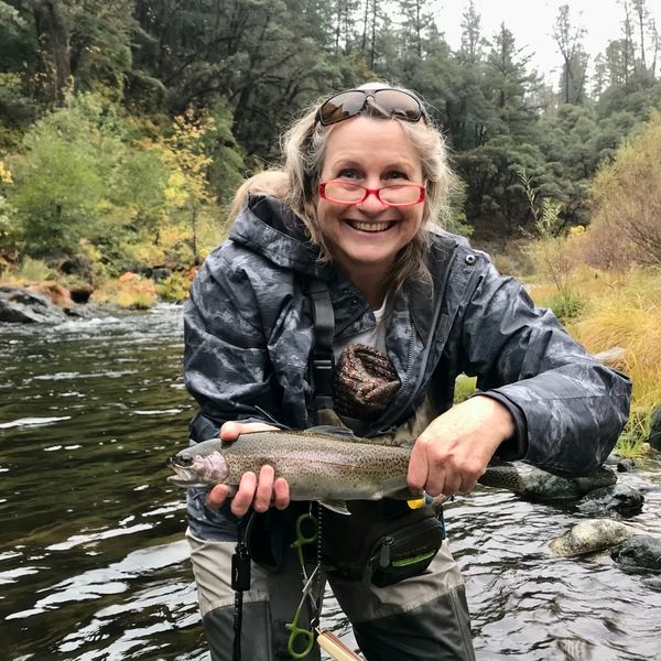 Smiling woman holding a freshly caught fish by a river in a forested area.
