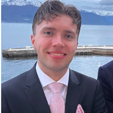 A young man in a suit smiling with a scenic water and mountain background.