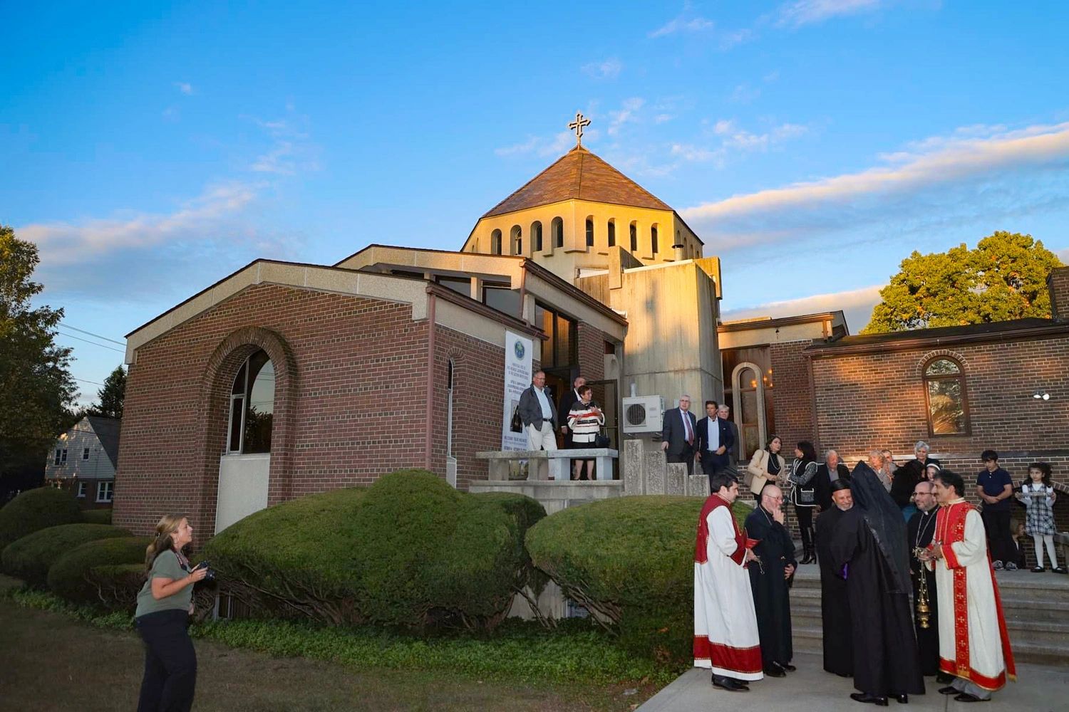 People gathered outside a church with clergy in ceremonial robes at sunset.