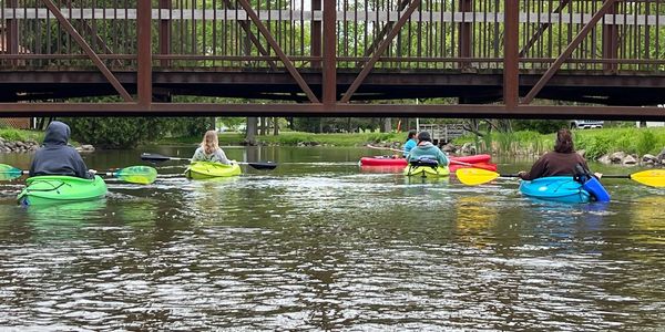 Group kayaking under a wooden pedestrian bridge on a calm river.