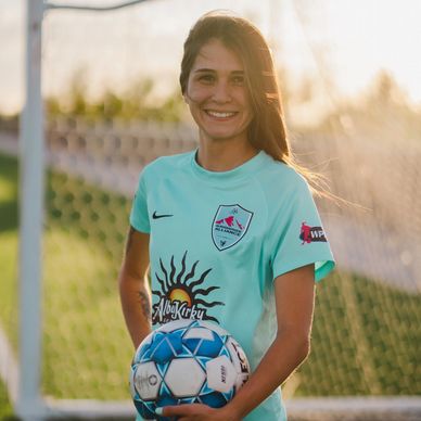 Female soccer player smiling with a ball in front of the goal.