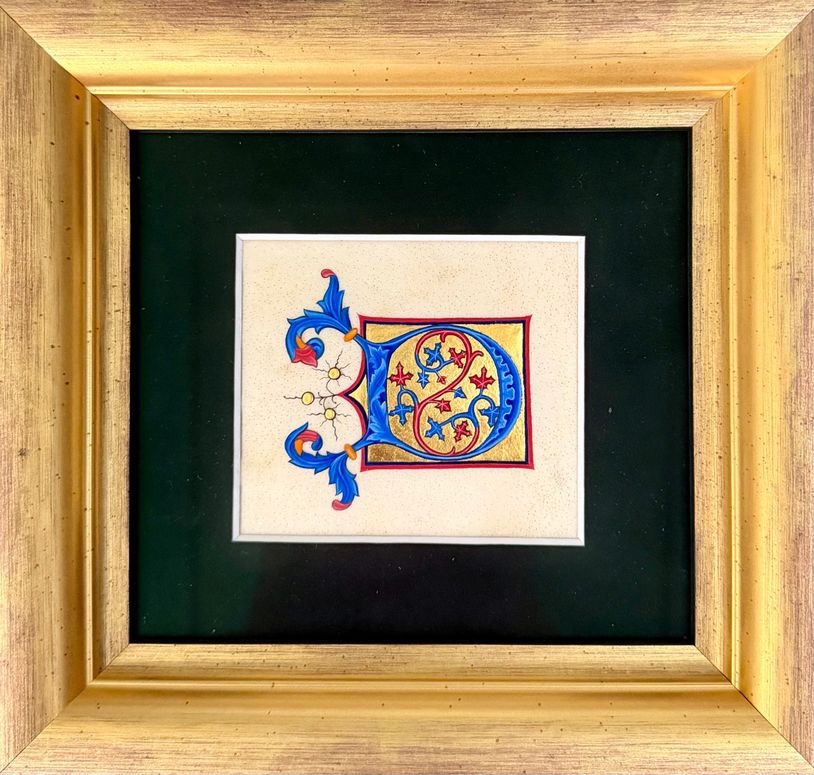 Ornate illuminated letter 'D' in blue and gold framed with a wooden frame.