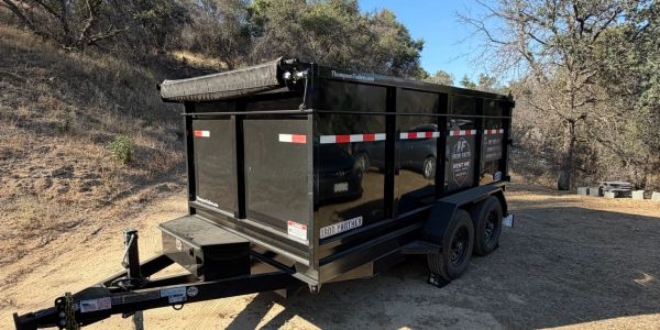 Black utility trailer parked on dirt near wooded area.