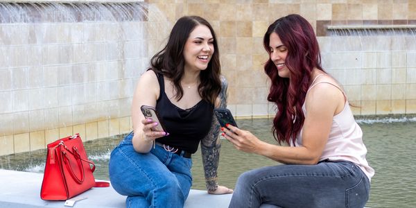 Two women sitting by a fountain, smiling and looking at their phones.