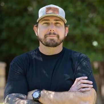 Man with beard wearing a cap and black shirt, arms crossed outdoors.