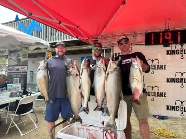 Three men proudly display large fish at a fishing competition under a red tent.