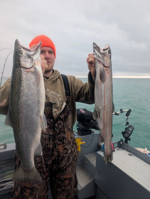 Man on boat holding two large fish, cloudy sky over water.