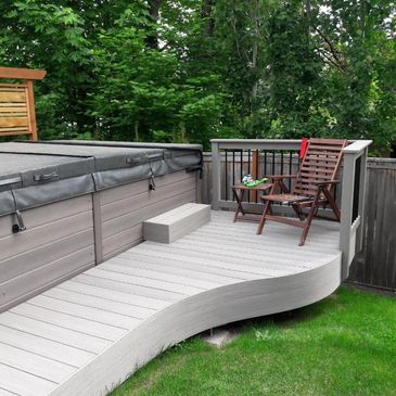 A deck with a curved finish and a hot tub. There is a folding chair on the deck.