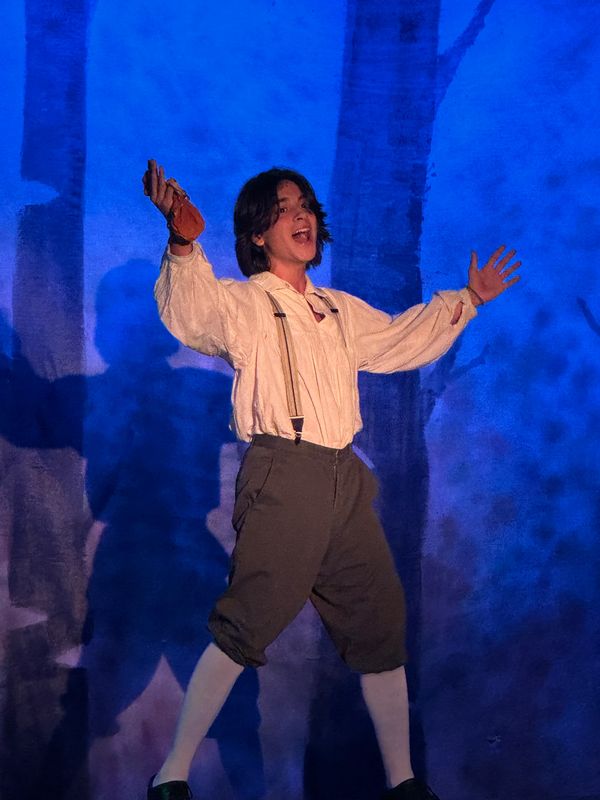 A performer joyfully gestures on stage with blue backdrop and theatrical costume.