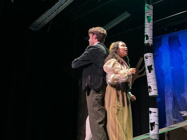 Two actors perform a dramatic scene on stage with a forest backdrop.