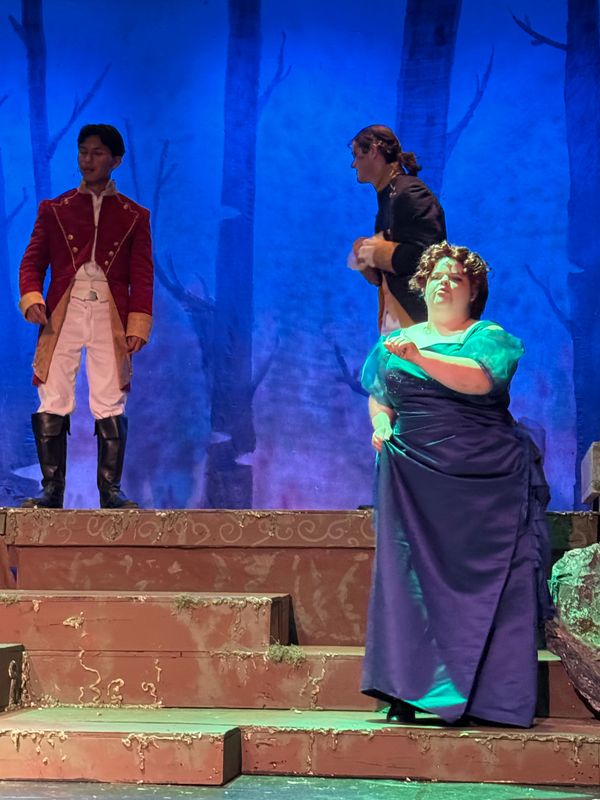 Actors performing a dramatic scene on a stage with a forest backdrop.