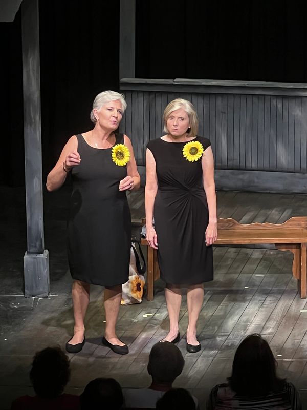 Two women in black dresses with sunflower pins perform on stage.