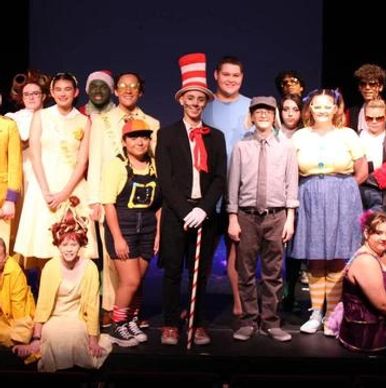 A diverse group in colorful costumes from a theatrical play poses on stage.