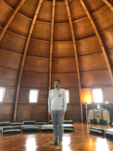 Man standing barefoot inside a wooden dome with circular seating.