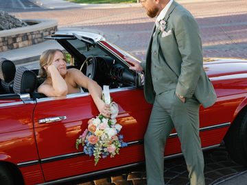 Bride in red car offering flowers to groom in green suit.