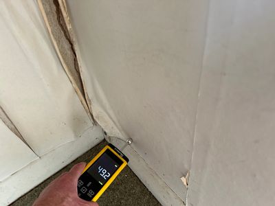 Hand holding moisture meter against damaged wall corner.