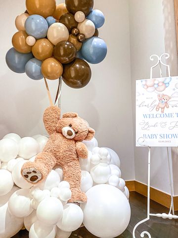 Teddy bear sitting on cloud of balloons, holding big balloon bouquet beside baby shower welcome sign