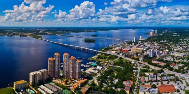 Aerial view of a coastal city with bridges and high-rise buildings under a partly cloudy sky.