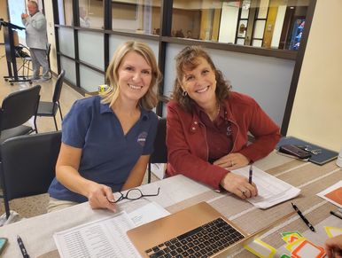 Two women smiling at a desk with papers and a laptop.