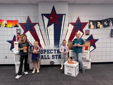 Four children stand in front of a wall with star decorations holding canned goods for a food drive.