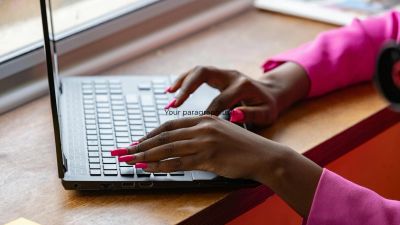 Person with pink nails typing on a laptop keyboard by a window.