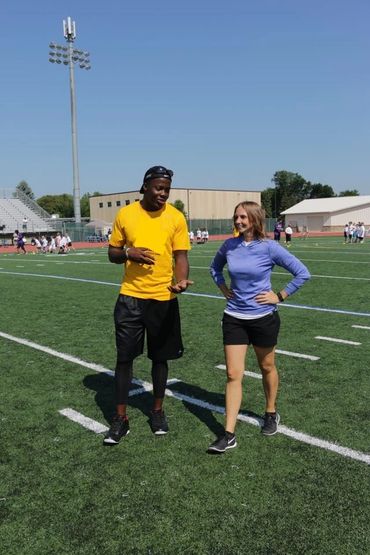 Two people walking and talking on a sports field under clear skies.
