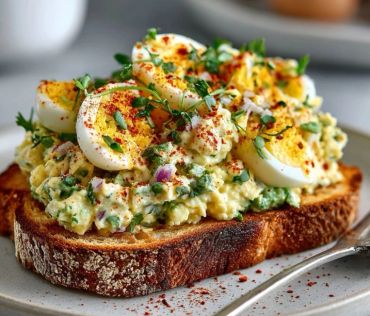 A slice of toast topped with creamy egg salad and sliced boiled eggs, garnished with herbs and paprika.