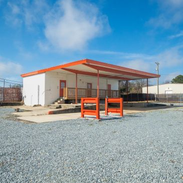 Small white building with orange roof in gravel lot under blue sky.