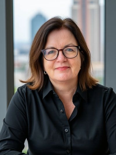 Photo portrait of the coach, Maureen Adams in a dark shirt by a window overlookiing a city landscape