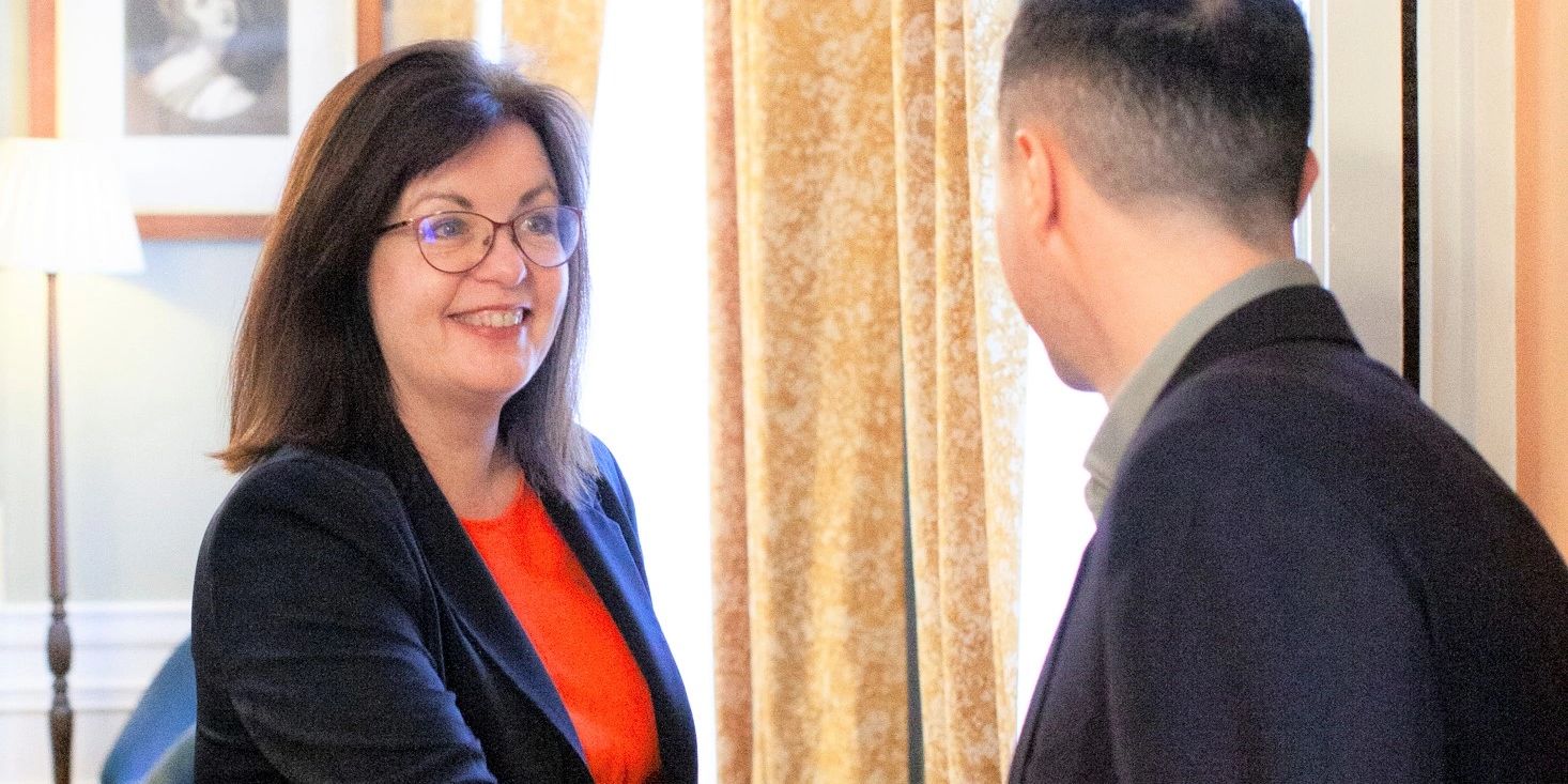 Woman in red dress and black jacket welcoming a man in blazer into a room and smiling.