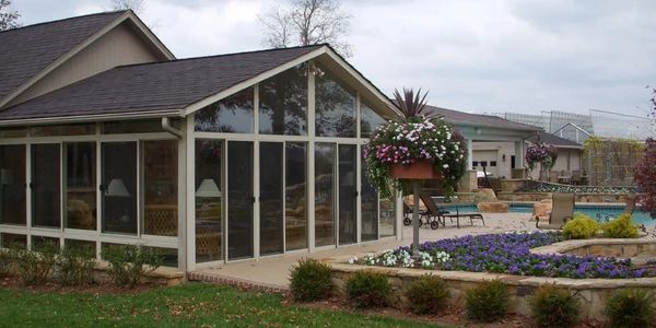 A glass sunroom attached to a house with a garden and pool in the background.