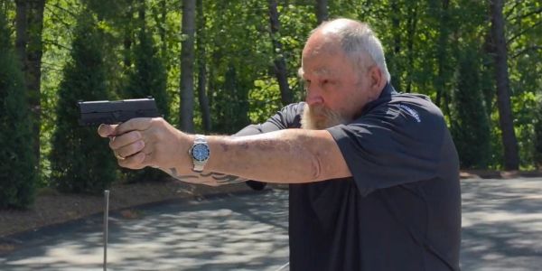 An elderly man aiming a handgun outdoors in a wooded area.