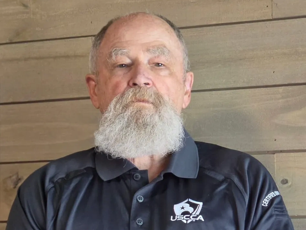 Bearded man wearing a black USCCA polo shirt against a wooden background.