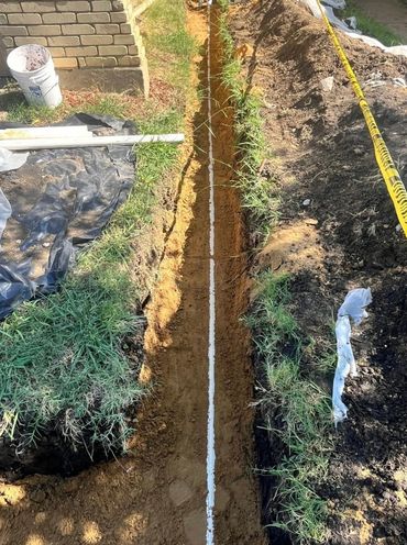 A narrow trench dug alongside a brick wall with a white pipe at the bottom.
