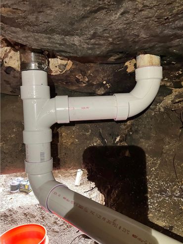 White PVC pipes connected with elbow and tee fittings in a basement or crawl space.