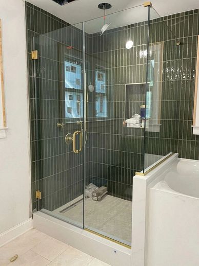Modern glass shower enclosure with green tiles and brass fixtures beside a white bathtub.