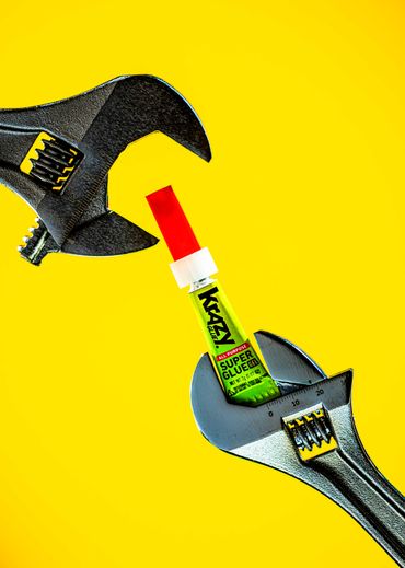Two adjustable wrenches gripping a tube of Krazy Super Glue against a yellow background.