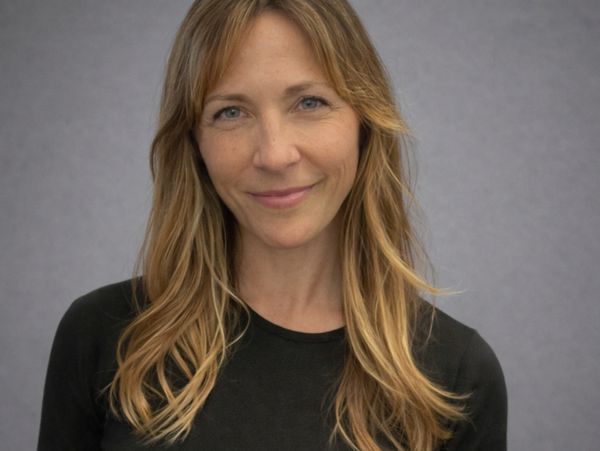 Smiling woman with long blonde hair wearing a black top against a gray background.
