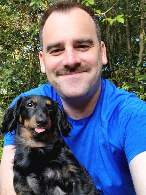 Man in blue shirt happily posing with a black and tan dog outdoors.